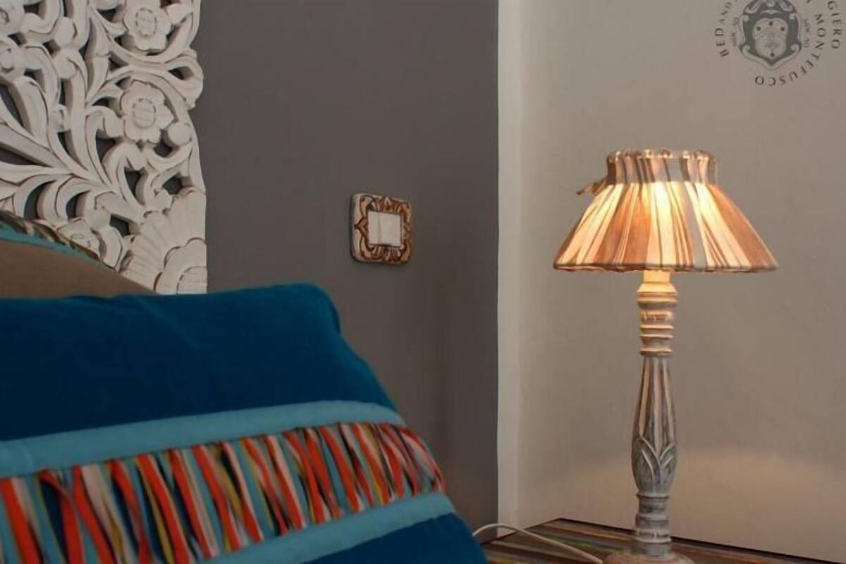 A bed with blue and multicolored pillows, a decorative wall panel, a wall-mounted light fixture, and a small framed object on the wall.