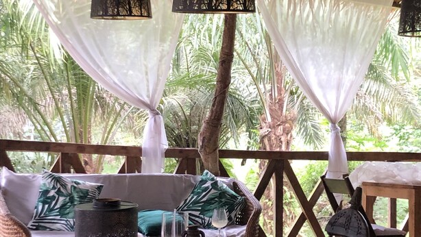 A covered outdoor seating area with wicker furniture, white cushions, and hanging lanterns. - Oveng Lodge (Nkolmétet)