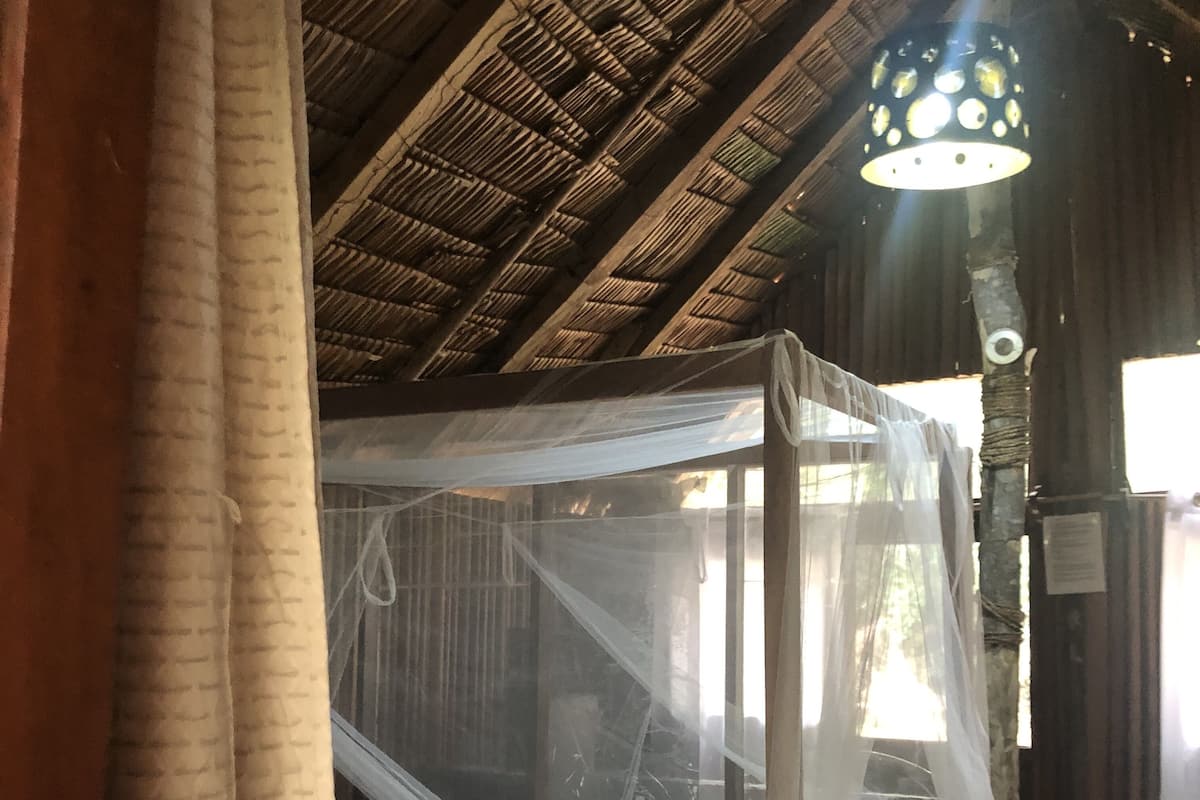A room with a bed under a mosquito net, wooden walls, a woven ceiling, and a hanging light fixture.
