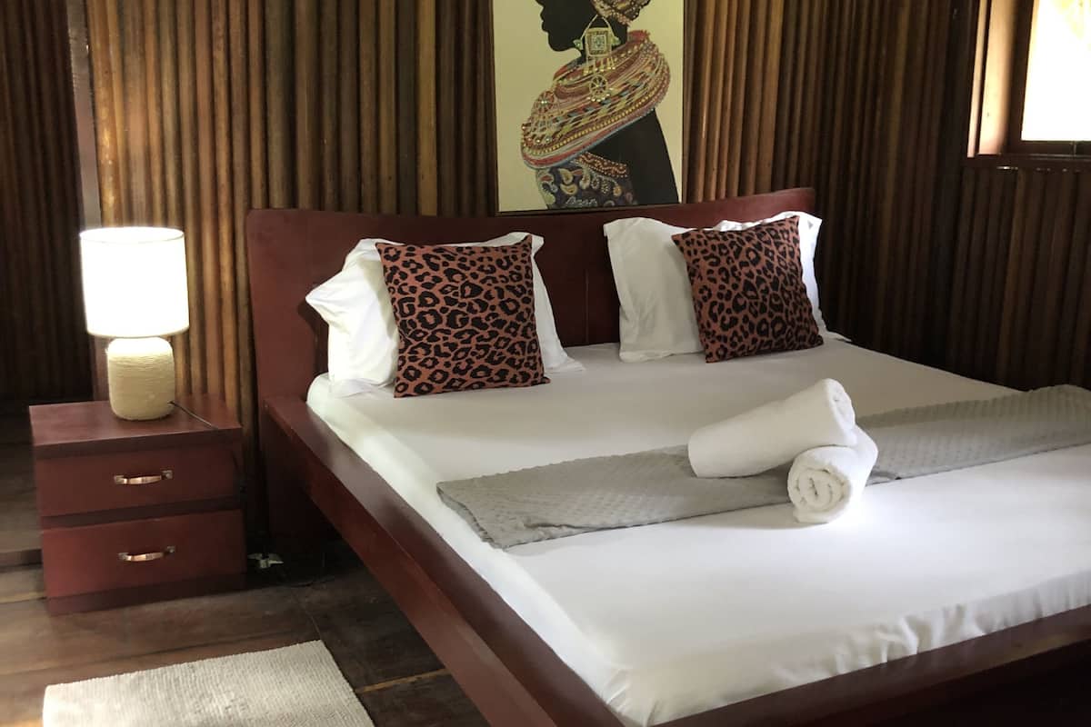 A neatly made bed with white linens and a rolled towel, a wooden nightstand with a lamp, and a patterned pillow.