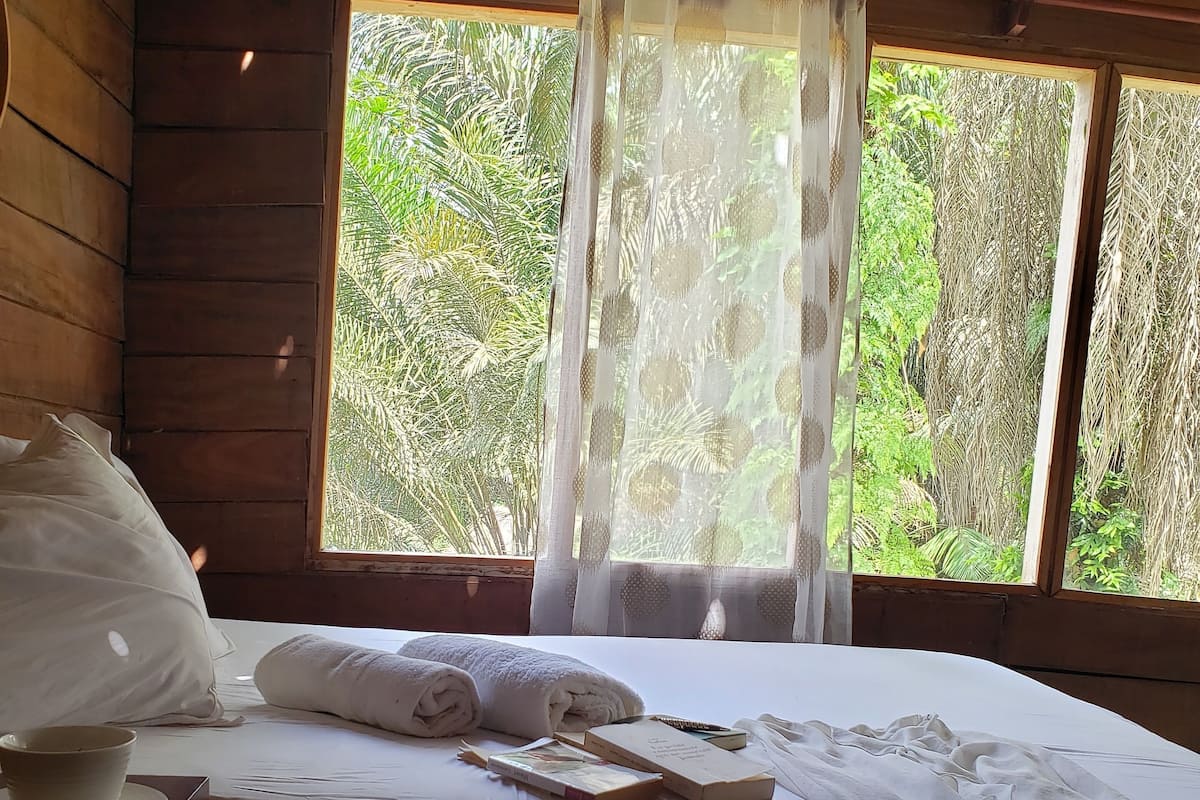 A wooden cabin room with a bed, a bedside table with a bowl, and a window with sheer curtains.