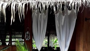 A wooden structure with a thatched roof and white curtains. There is a wooden staircase leading to an open area with a large green plant. - Oveng Lodge (Nkolmétet)