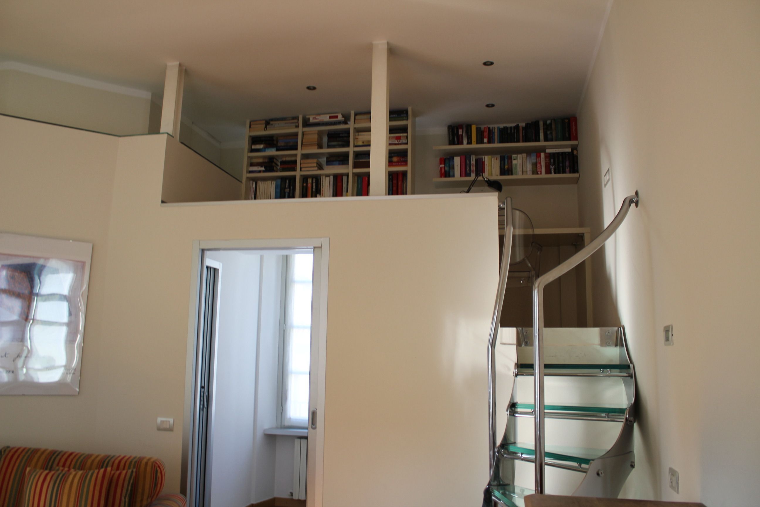 Amplia sala de estar con techo alto, una escalera con barandillas de vidrio y una estantería llena de libros.