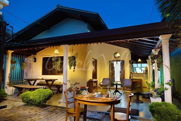 A covered outdoor seating area with tables and chairs, a small garden, and a building entrance.