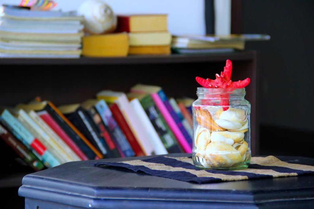 Un frasco de vidrio con conchas marinas y un adorno rojo sobre una mesa cubierta con un mantel a rayas, frente a una estantería llena de libros.