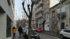 Una estrecha calle urbana bordeada de edificios de varios pisos, coches aparcados y algunos peatones.