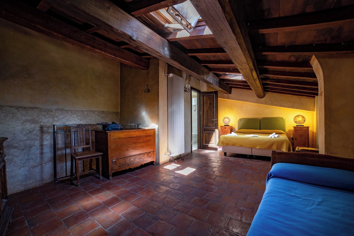 A rustic bedroom with exposed wooden beams, a brick floor, a bed with a yellow bedspread, a wooden dresser, and a small table with a lamp.