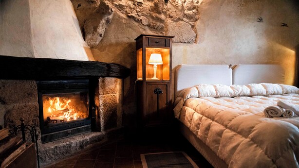 Una habitación acogedora con chimenea de piedra, una cama con edredón blanco y una mesita de noche con lámpara.