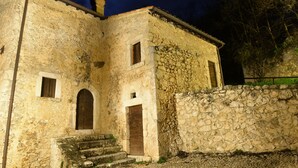 Un edificio de piedra con una puerta y ventana de madera, un conjunto de escalones que conducen a la entrada y un muro de piedra a la derecha.