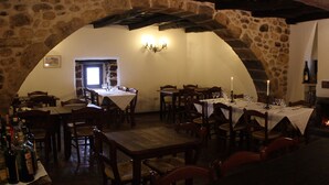 Un comedor rústico con paredes de piedra, mesas y sillas de madera. Hay una chimenea encendida y una estantería con libros y una espada.