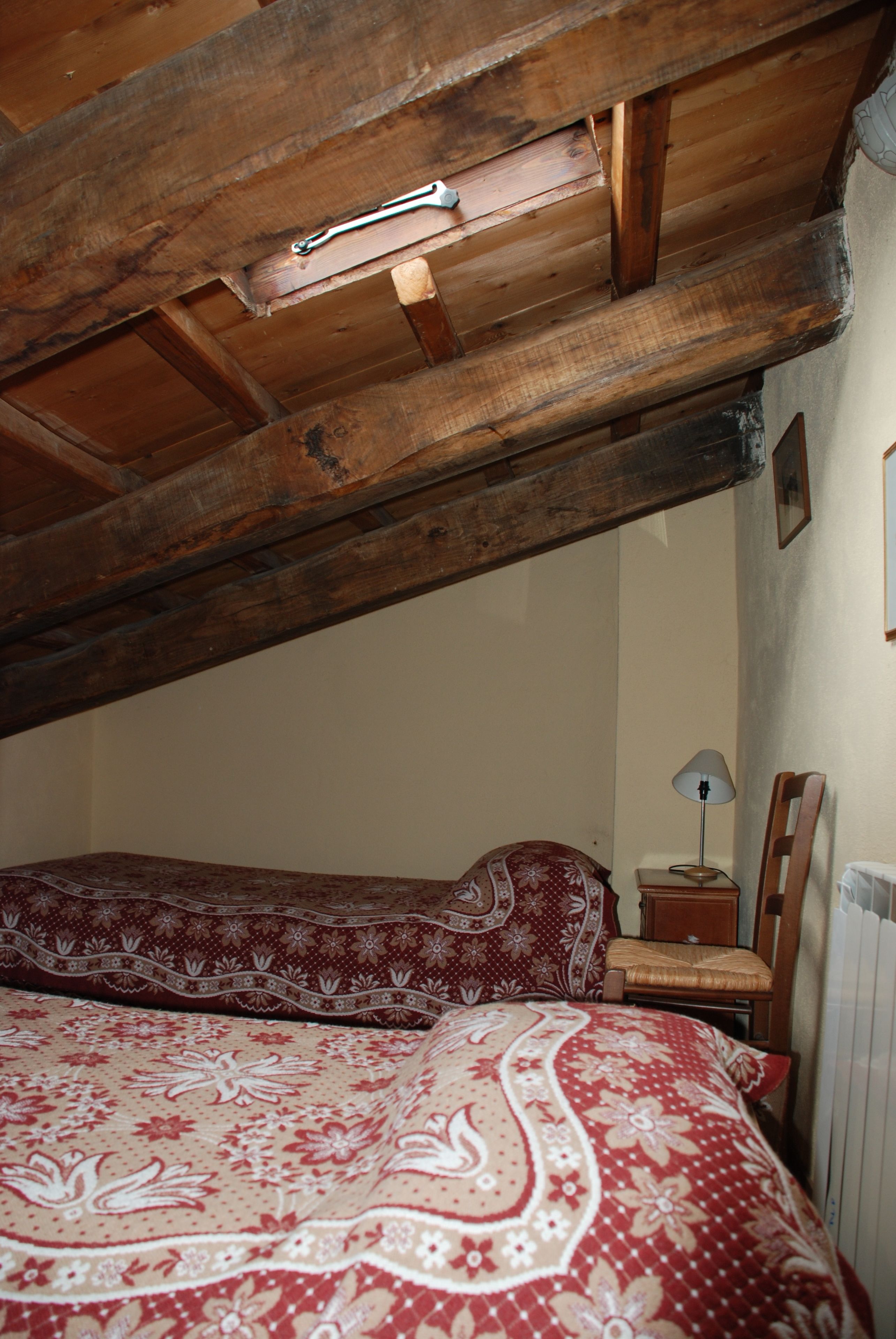 A room with a sloped wooden ceiling, two beds with patterned bedding, a small bedside table with a lamp, and a chair.
