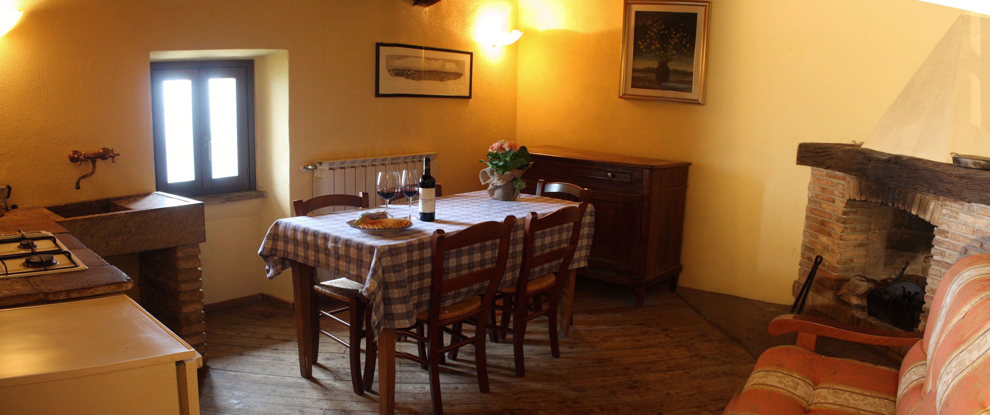 Un coin repas chaleureux avec une table dressée pour deux personnes, des chaises en bois, un petit vase avec des fleurs et une cheminée en briques.