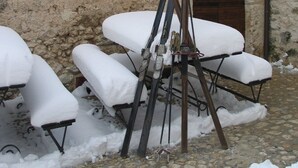 Un edificio de piedra rústica con una puerta de madera, un banco cubierto de nieve y esquíes apoyados en la pared.