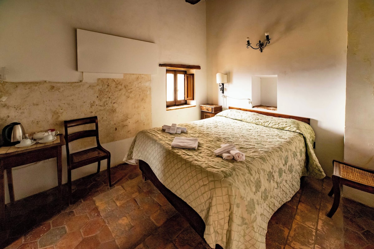 A bedroom with a bed, a chair, a table with a kettle, and a window.