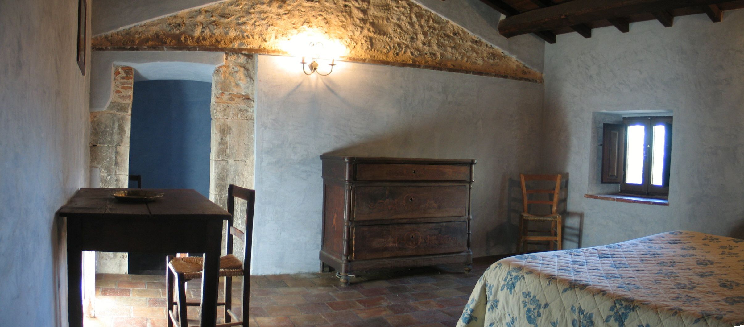 A rustic room with a stone ceiling, a wooden dresser, a bed with a floral bedspread, a chair, and a small window.