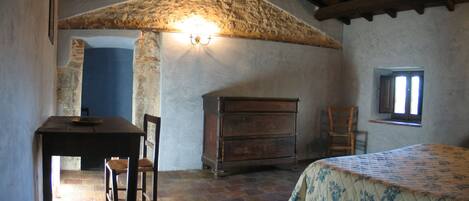 Una habitación rústica con techo de piedra, un aparador de madera, una cama con colcha estampada, una silla y una ventana pequeña.