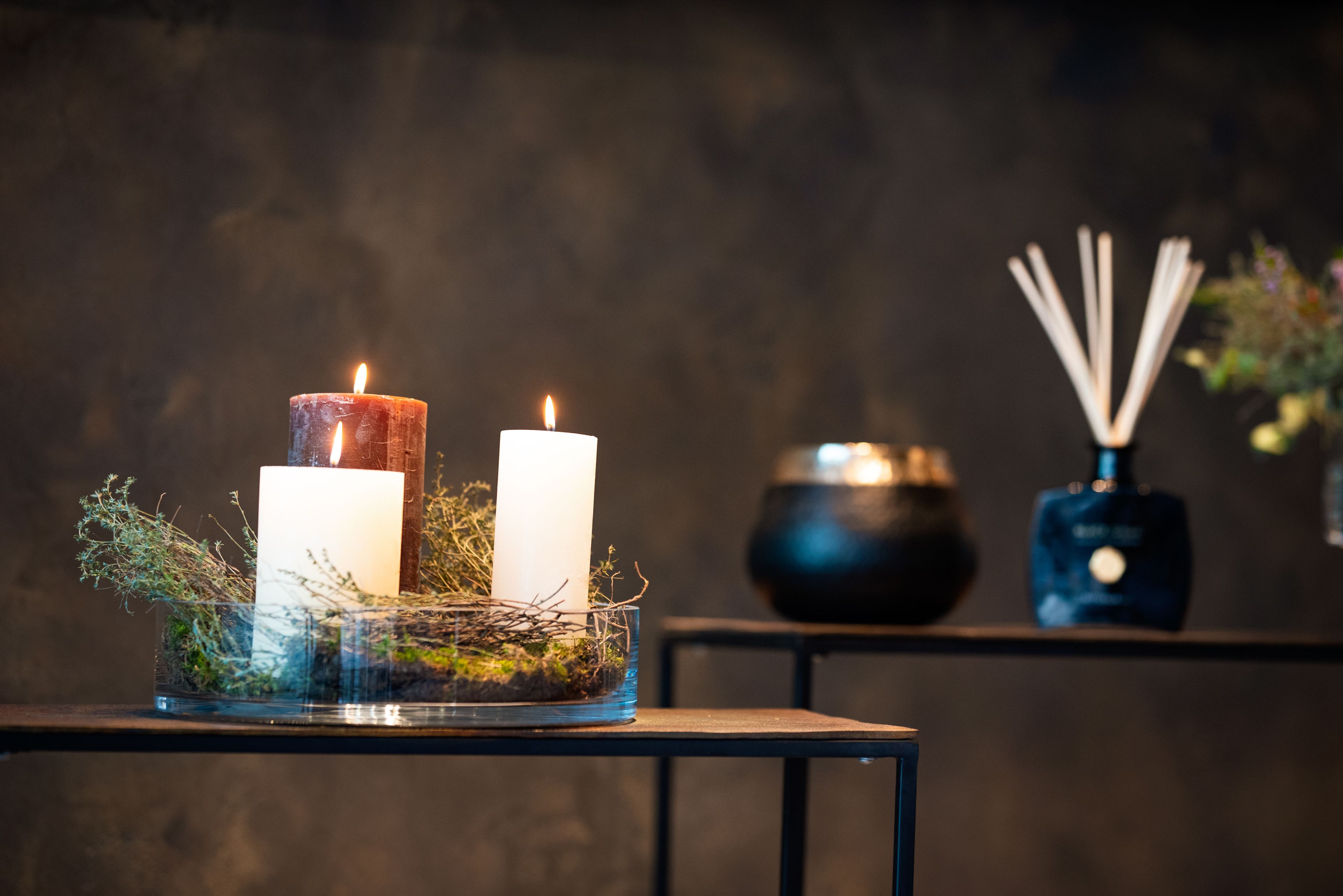 Una mesa con dos velas encendidas en un recipiente de vidrio rodeada de follaje.