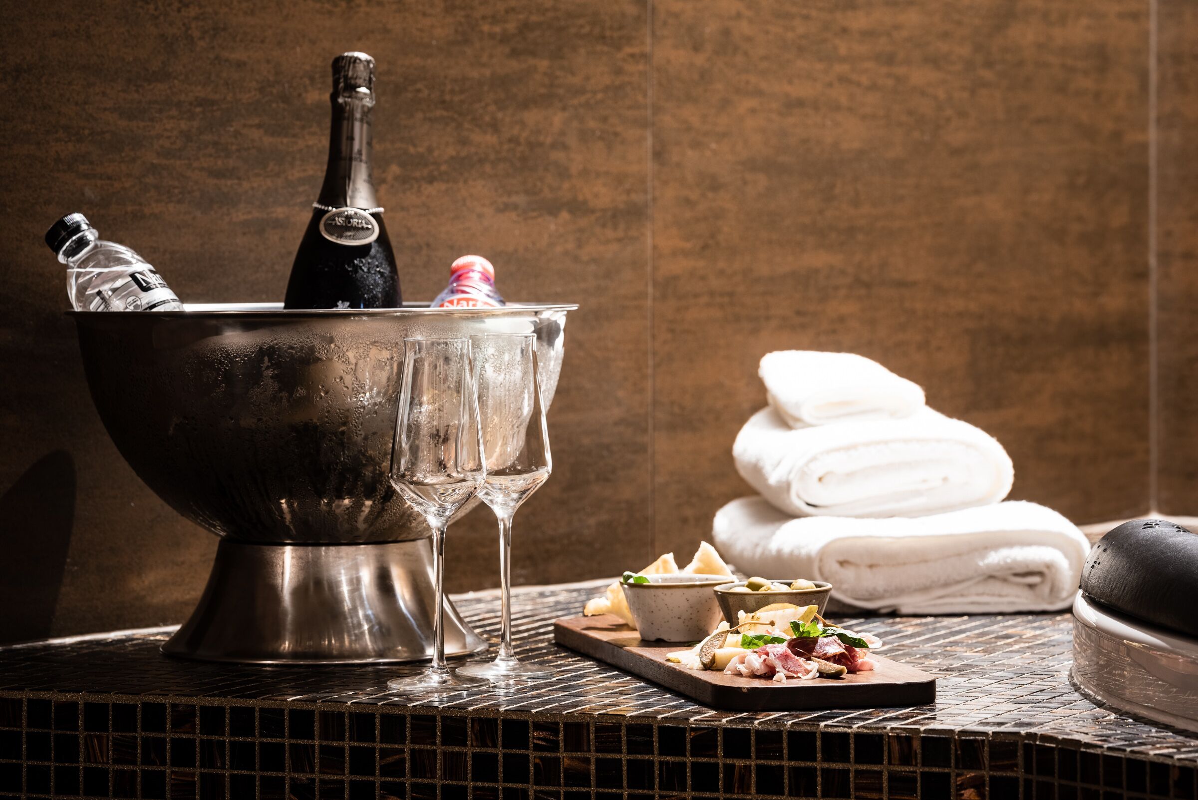A metallic bucket with a bottle and glasses, a tray with cheese and olives, and folded towels against a textured wall.