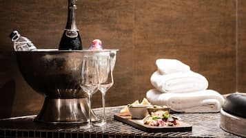 A metallic bucket with a bottle and glasses, a tray with cheese and olives, and folded towels against a textured wall.
