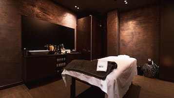 A modern bathroom with a large mirror, dark wood paneling, a sink, and a massage table with towels.