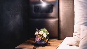 A modern hotel room with a wooden nightstand, a wall-mounted lamp, and a floral arrangement.