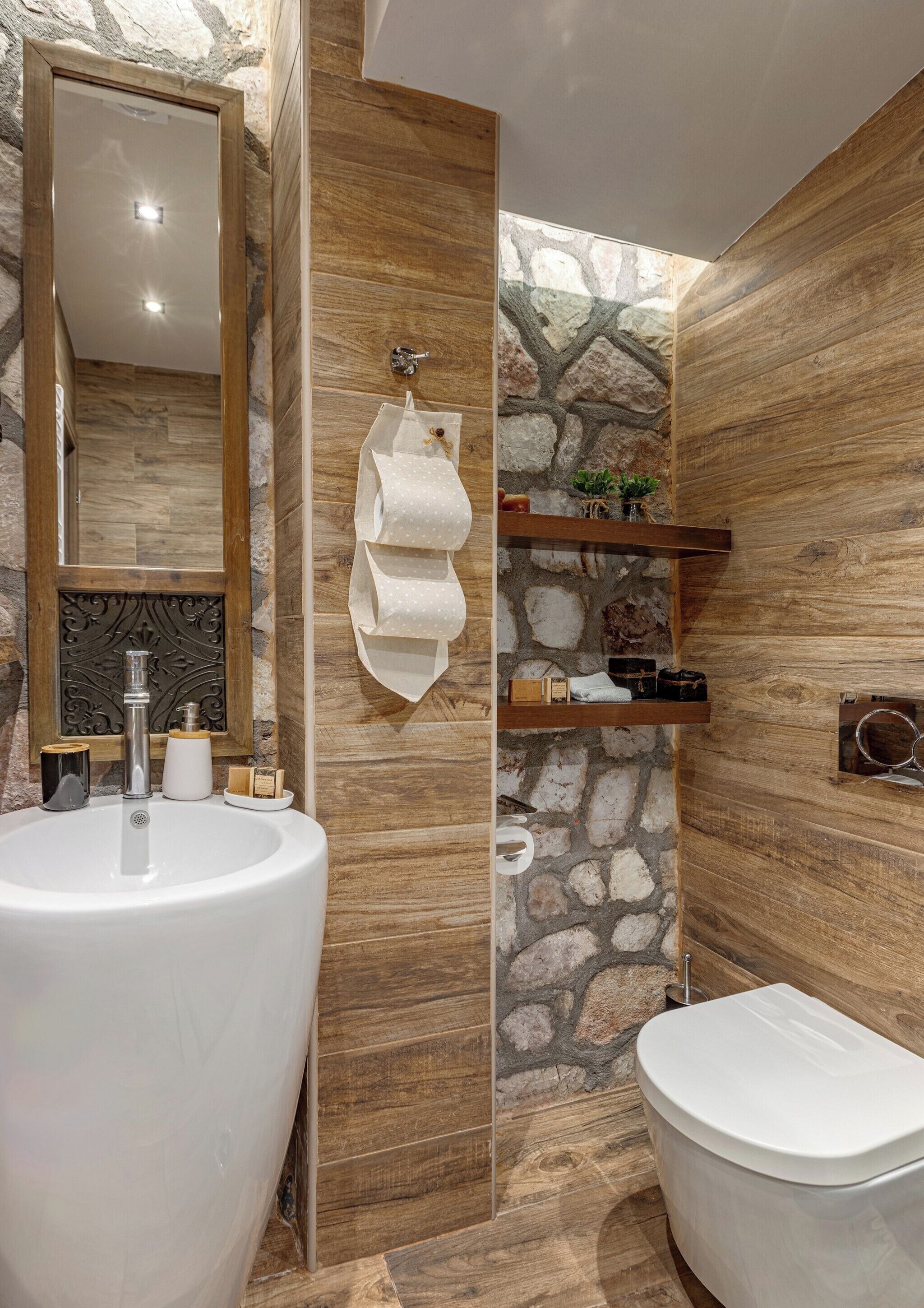 Un baño con un lavamanos grande, inodoro y espejo. Hay estantes con artículos de aseo y un perchero para toallas.