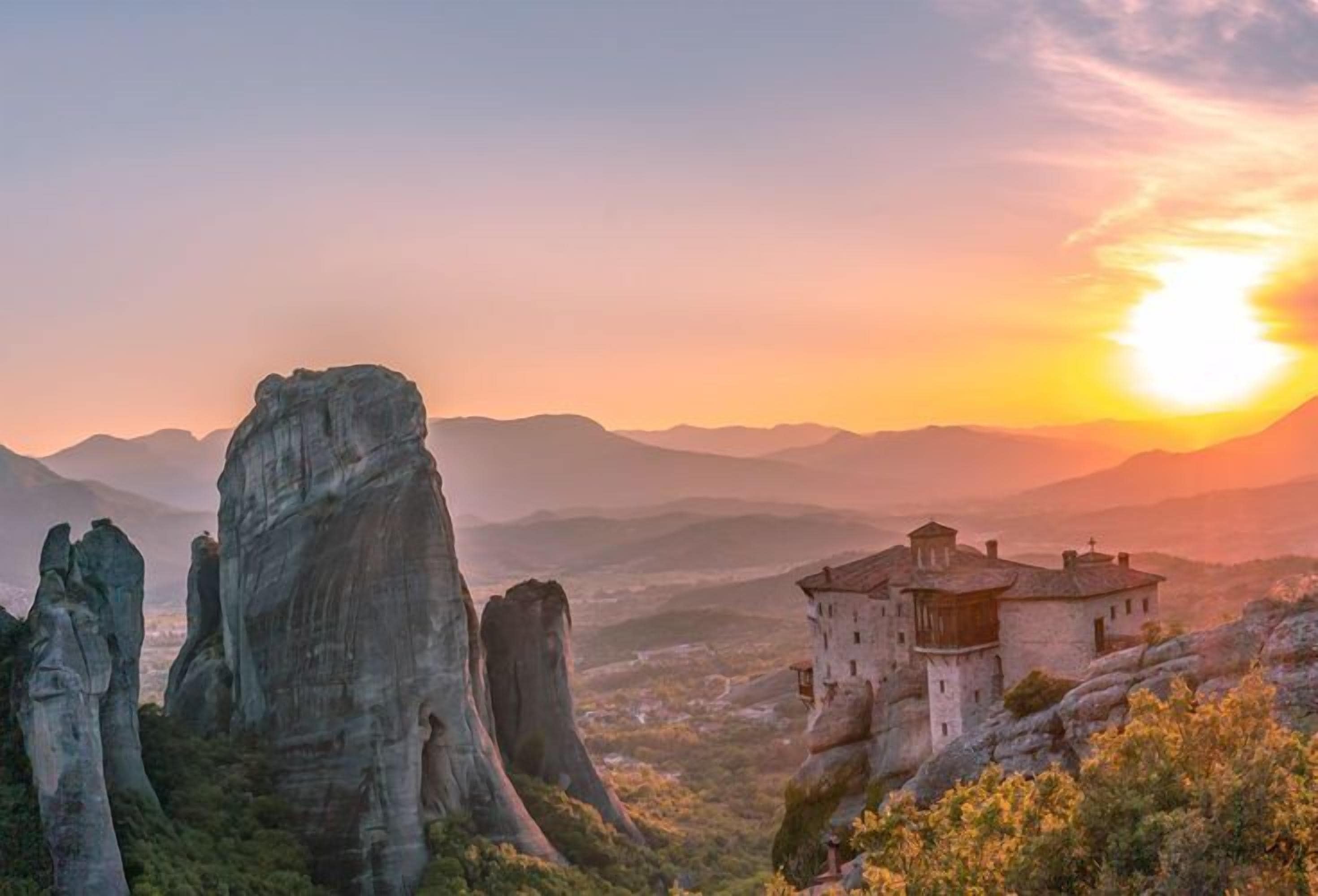 A historic stone building with a prominent tower, surrounded by rocky terrain and a sunset in the background.