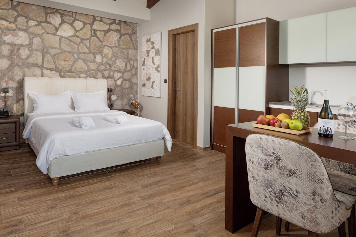 Ein Schlafzimmer mit Steinwand, einem Bett mit weißen Leinen, einem Holztisch mit Obstschale, einem Stuhl und einer Minibar.