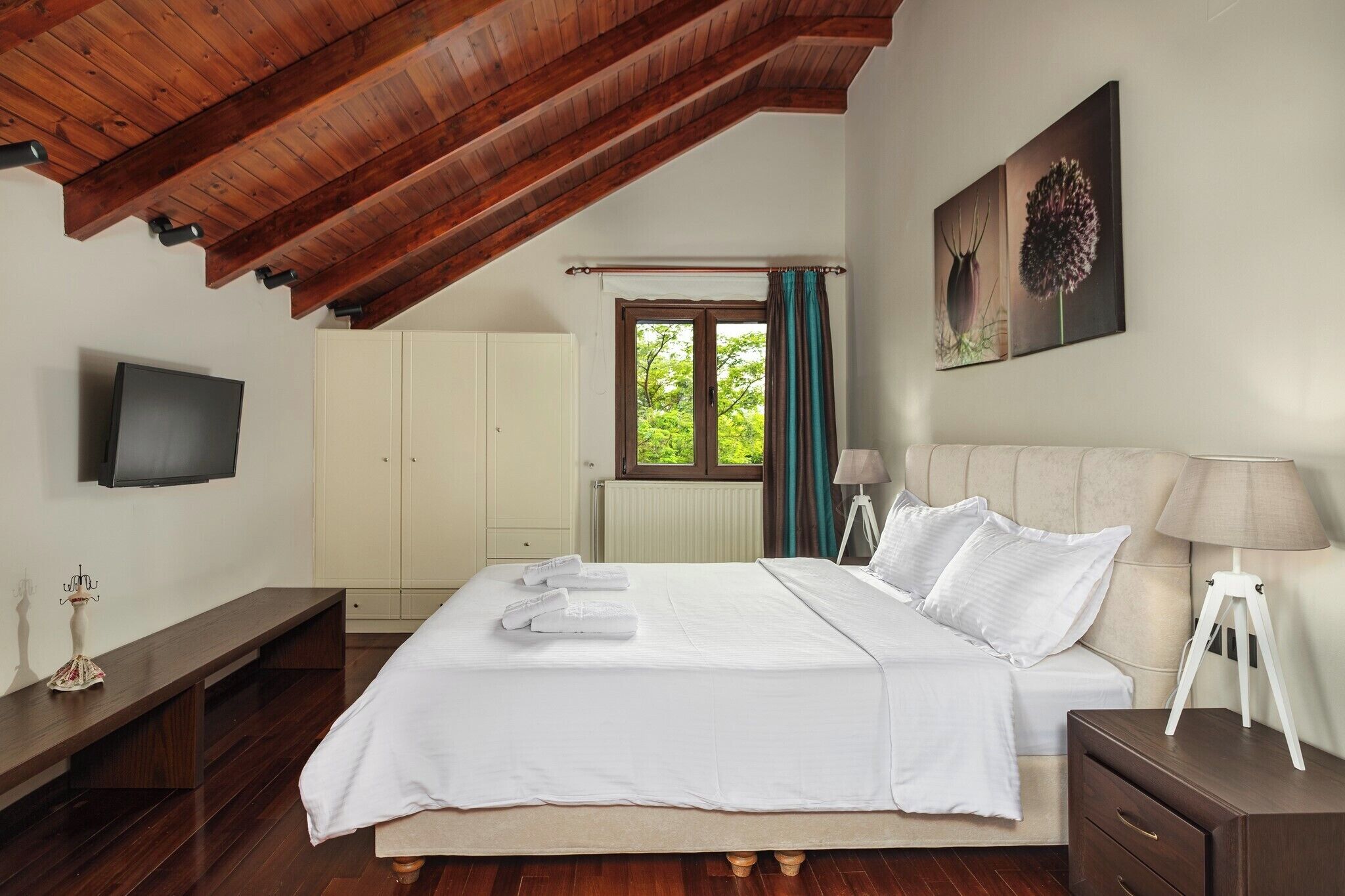 Un dormitorio con techo de madera, una cama con sábanas blancas, una lámpara de noche y una ventana con cortinas.