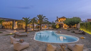A pool area with lounge chairs, a stone patio, and a building with large windows and outdoor lighting. - Vintage Suites Trikala (Trikala)