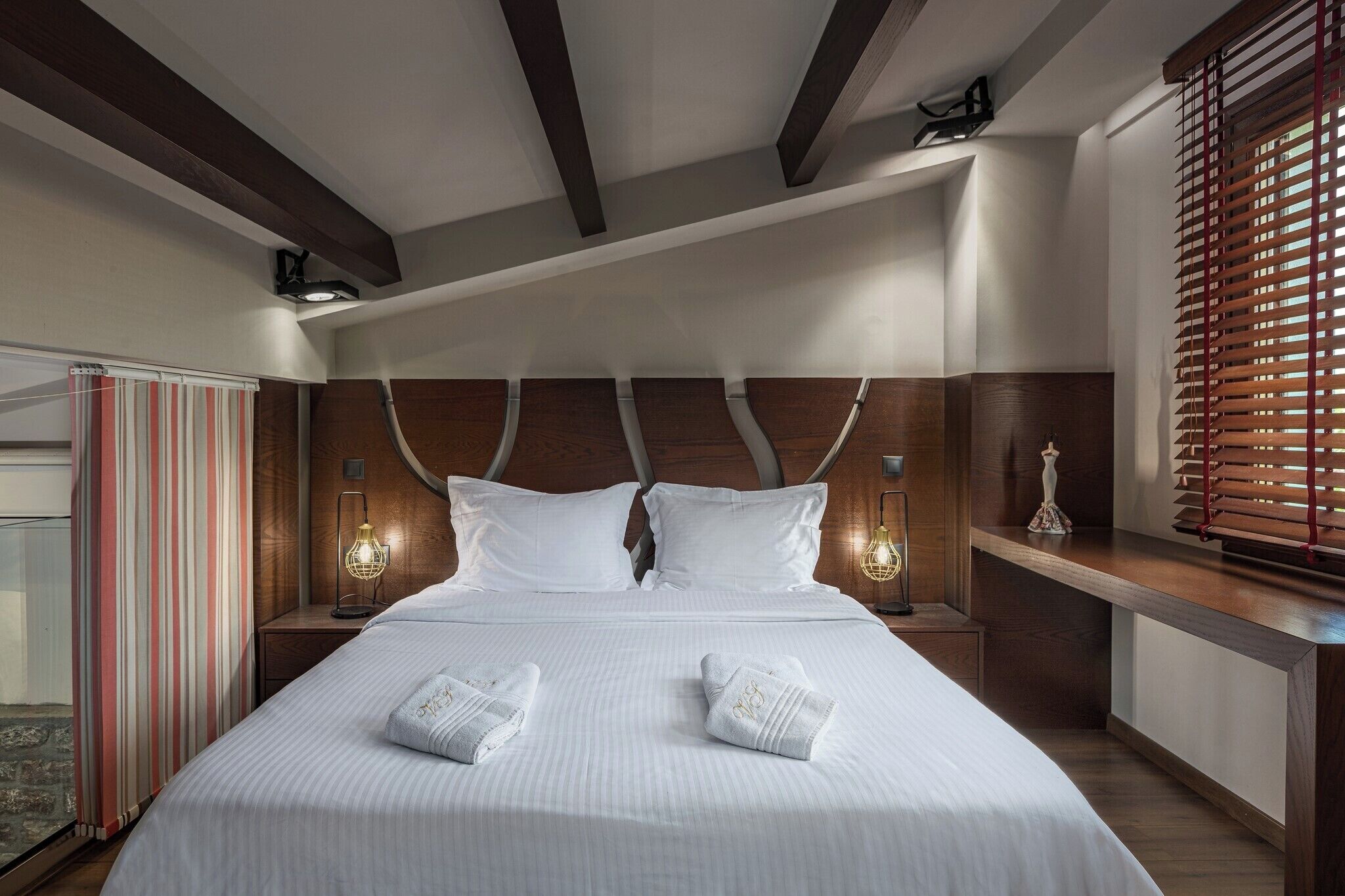 Una habitación de hotel con una cama grande, cabecera de madera, dos lámparas de noche y una ventana con persianas.