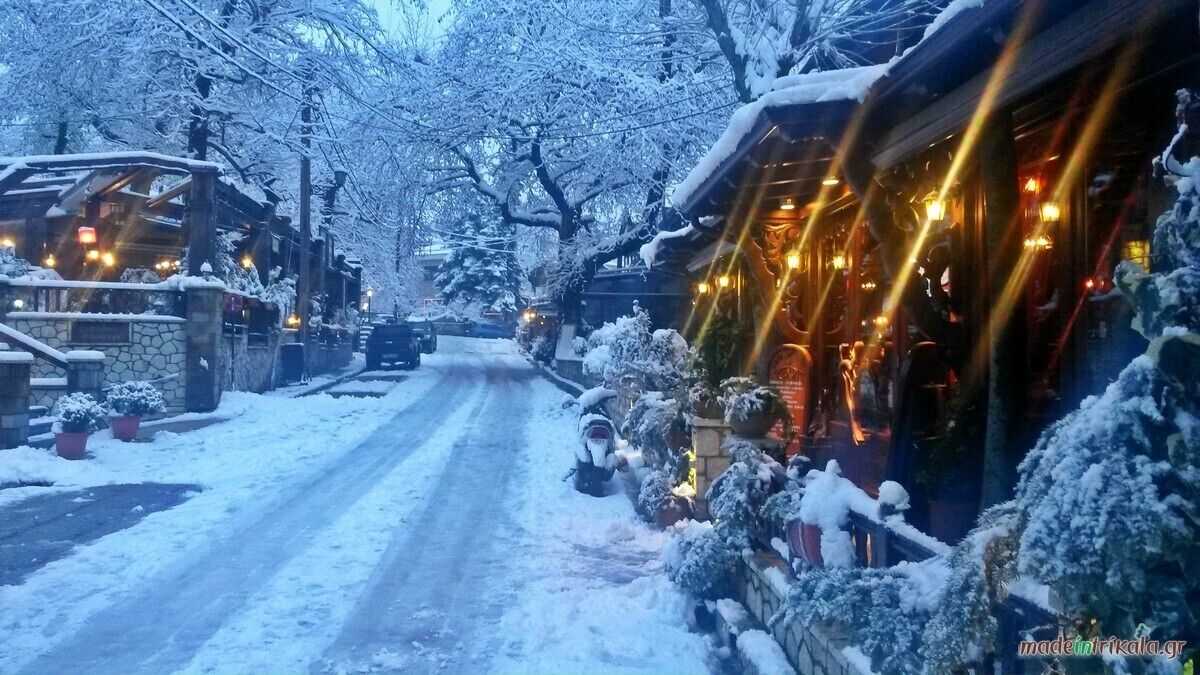 A snow-covered street with buildings on either side, including a restaurant with outdoor seating and lights.