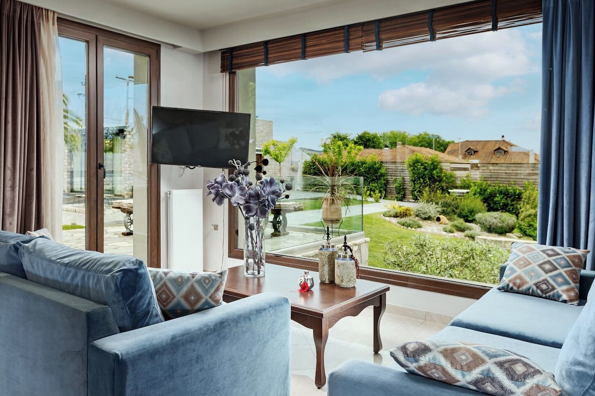 Ein modernes Wohnzimmer mit großem Fenster, einem Flachbildfernseher und Blick auf einen Garten sowie Gebäude.