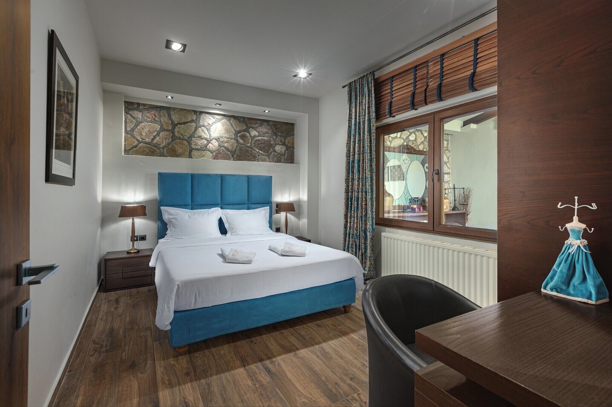 A hotel room with a bed, bedside tables, a dresser, a window with curtains, and a small table with a dress on it.