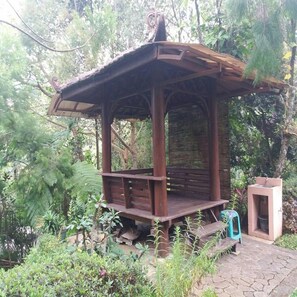 Un quiosco de madera con techo inclinado, rodeado de abundante follaje y un sendero de piedra.