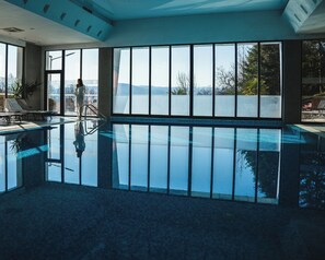 A large indoor swimming pool with clear glass walls, a lounge area with chairs, and a view of the outdoors. - Medite Spa Resort & Villas (Sandanski)