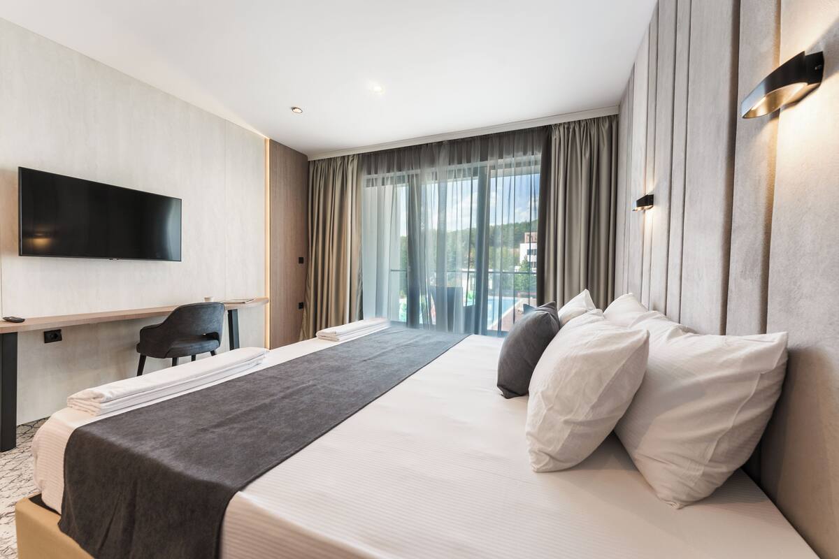 A modern hotel room with a large bed, a flat-screen TV, a desk, and a view of the outdoors through large windows.