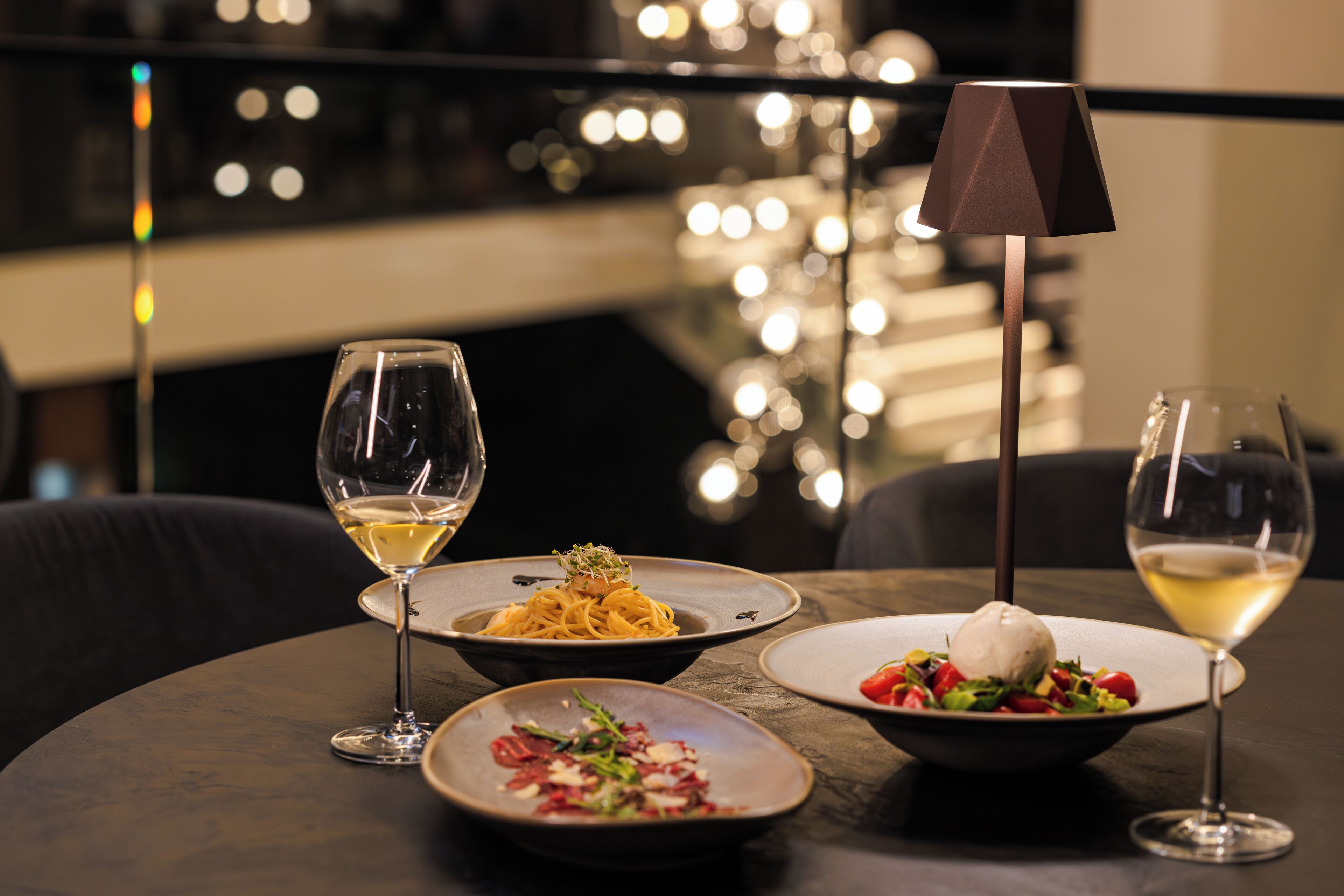 A table with three dishes and two glasses of white wine, set against a dark background with blurred lights.