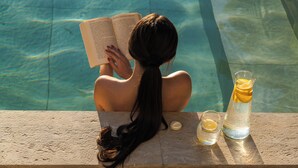 Une personne est assise au bord d’une piscine, en train de lire un livre, avec un verre d’eau et une tranche de citron.