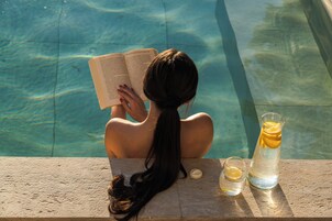 Una persona leyendo un libro junto a una piscina, con un vaso de agua y una rodaja de limón.