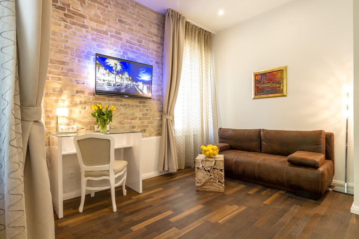 A living room with a brick wall, a TV mounted above a desk, a brown leather sofa, and a small wooden side table with a vase of yellow flowers.