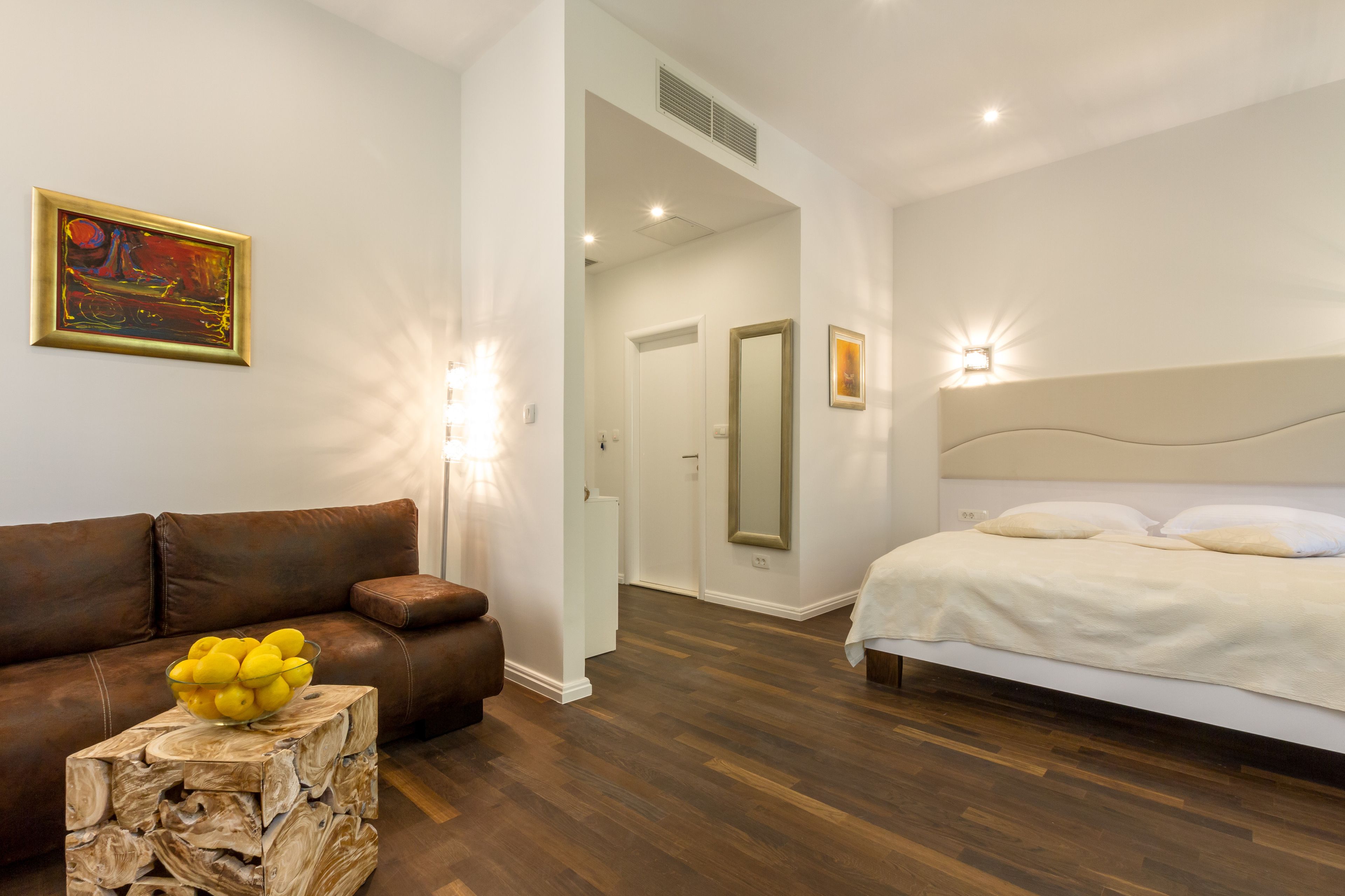 A modern bedroom with a bed, a brown leather sofa, a wooden side table with a bowl of lemons, and a painting on the wall.