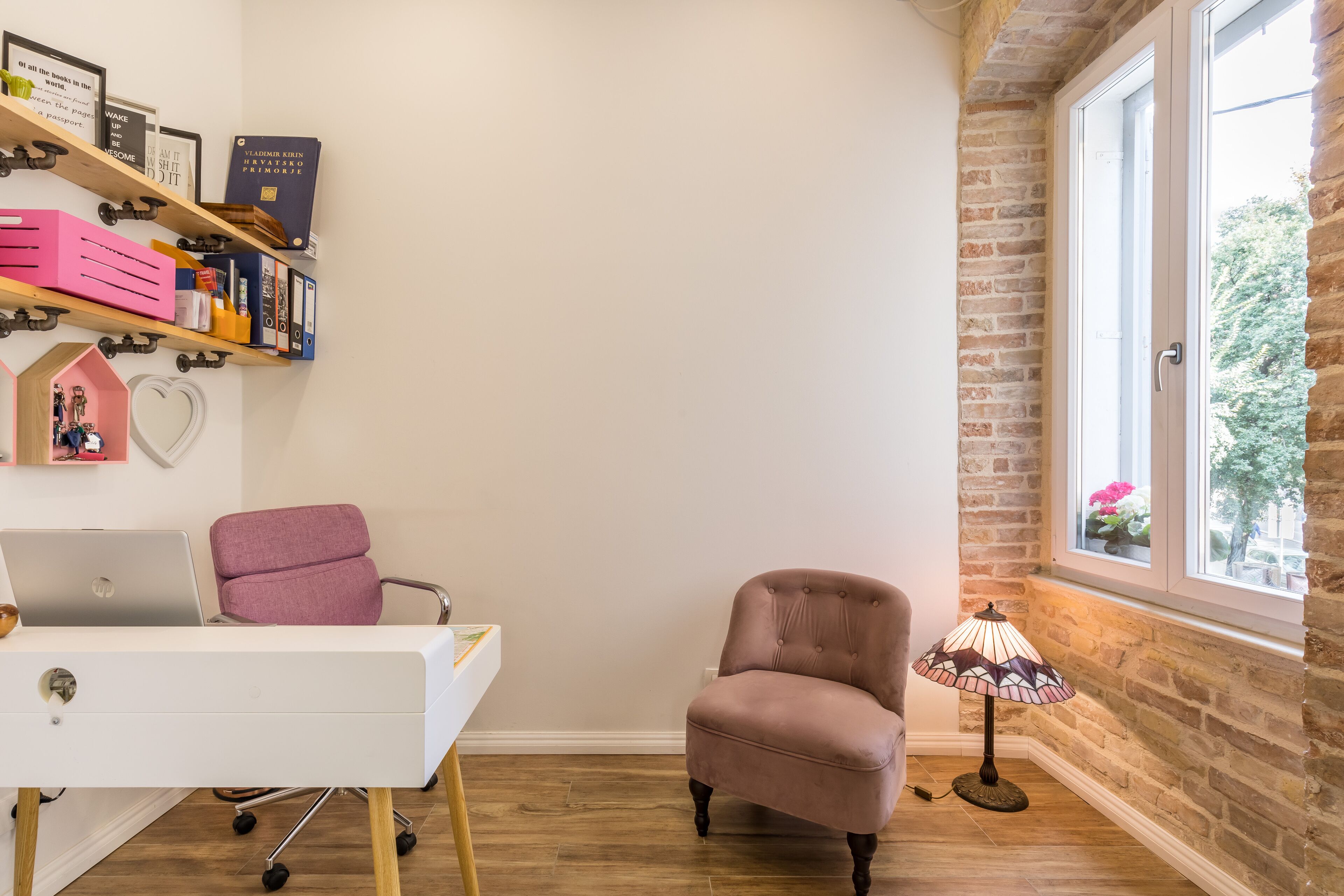 A room with a white desk, a pink chair, a bookshelf with books, and a lamp near a window.