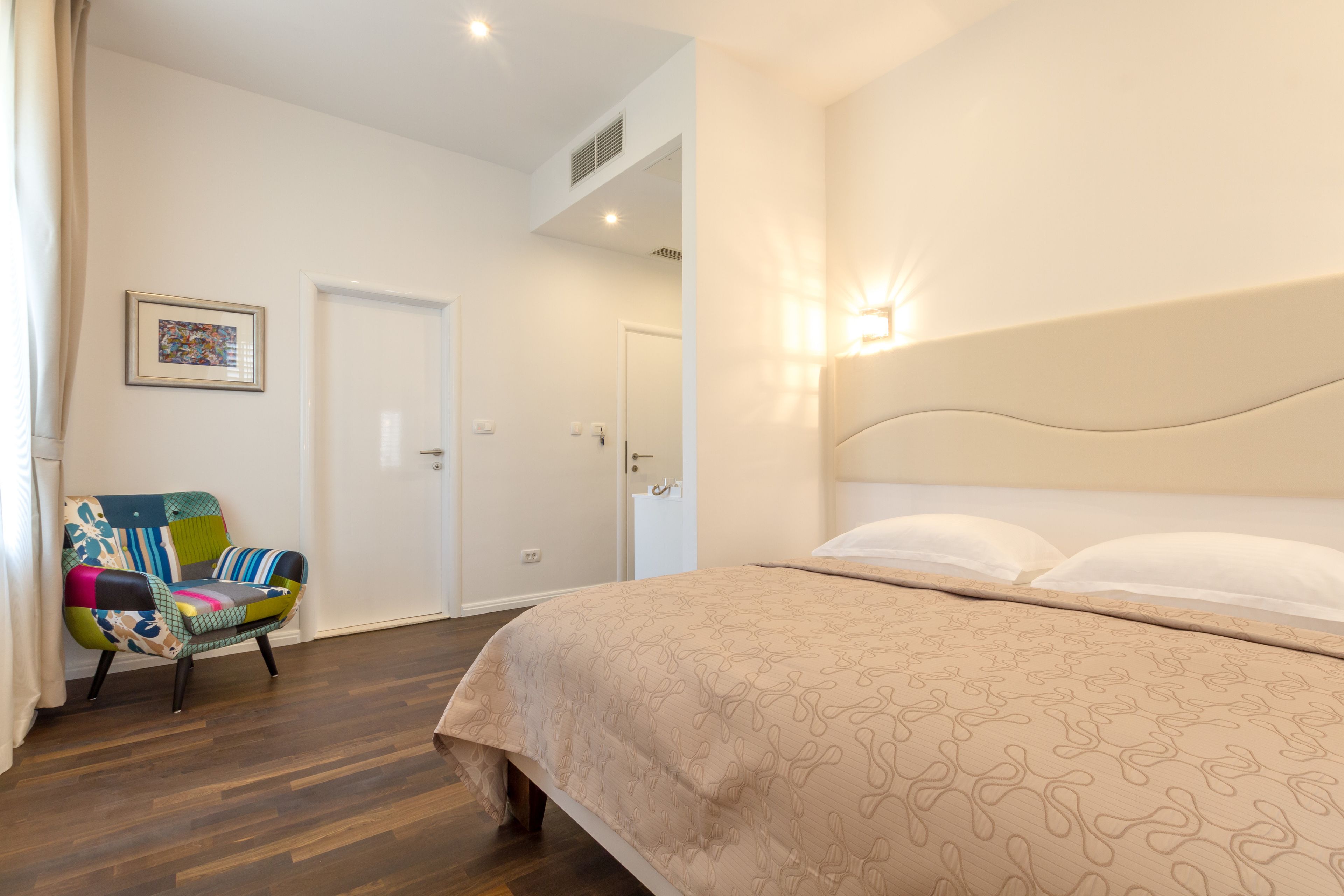 Een slaapkamer met een bed, een kleurrijke stoel, een deur en een wandlamp.