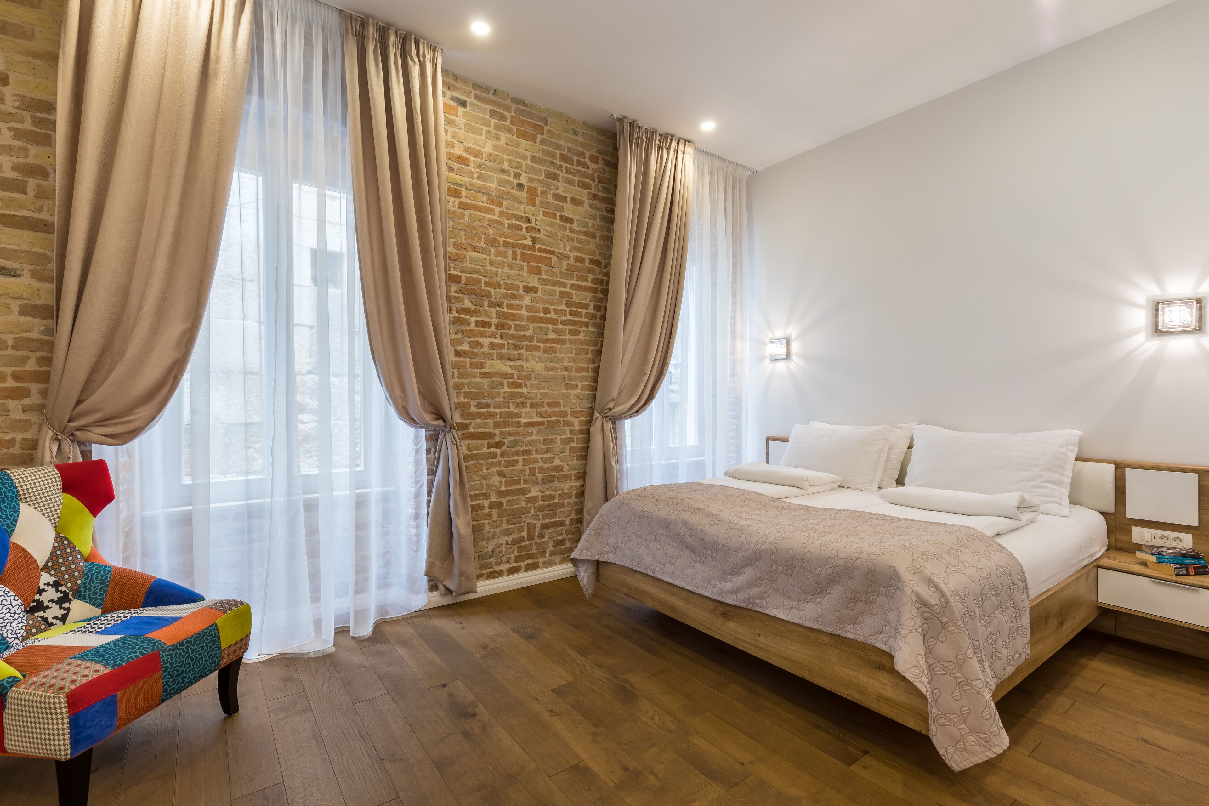A bedroom with a wooden bed, a colorful armchair, and a brick wall.