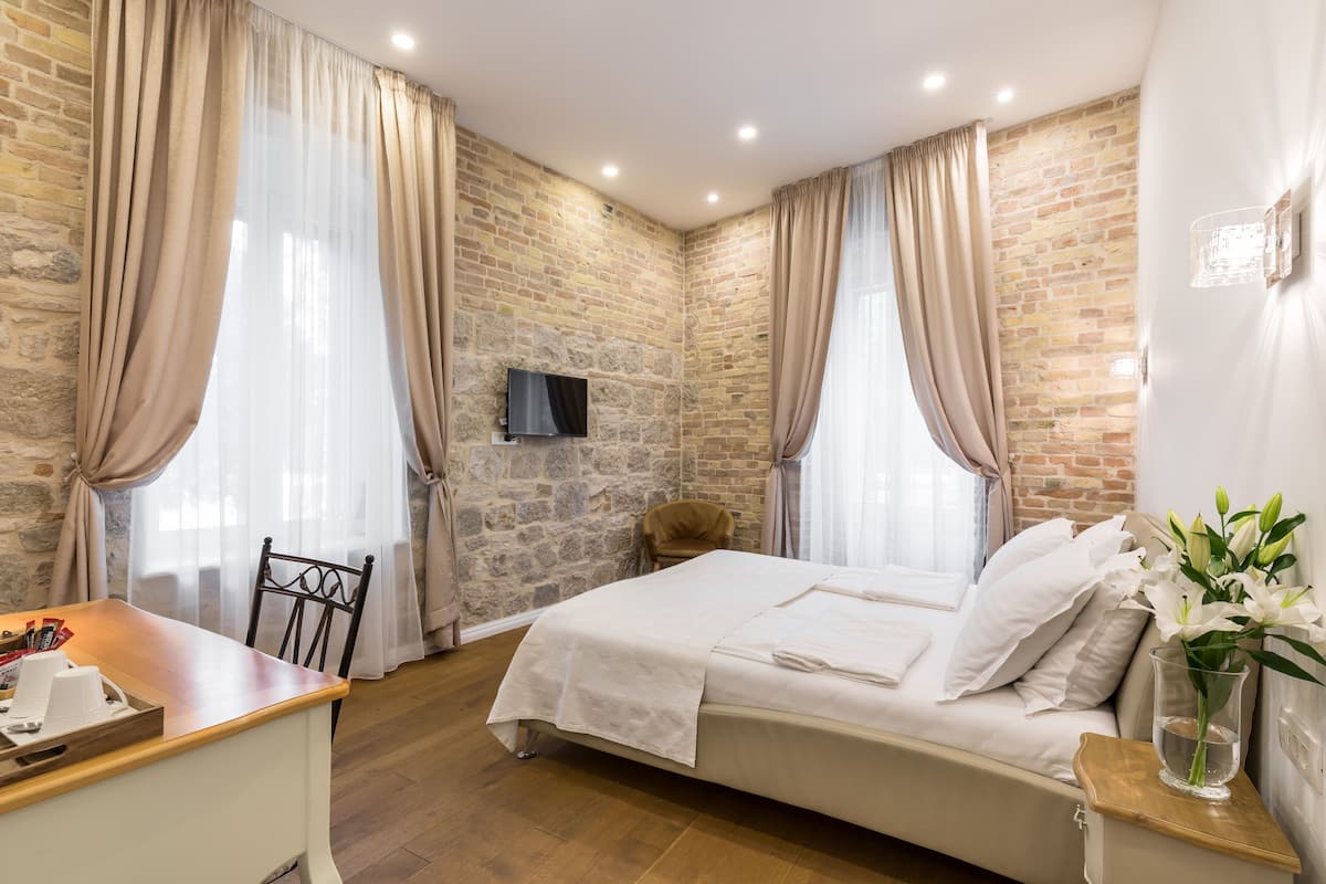 A bedroom with a stone wall, a bed with white linens, a desk with a chair, and a TV mounted on the wall.