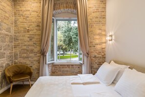 A bedroom with a brick wall, a window with curtains, a bed with white linens, and a wooden chair. - Pellegrini Luxury Rooms (Split)
