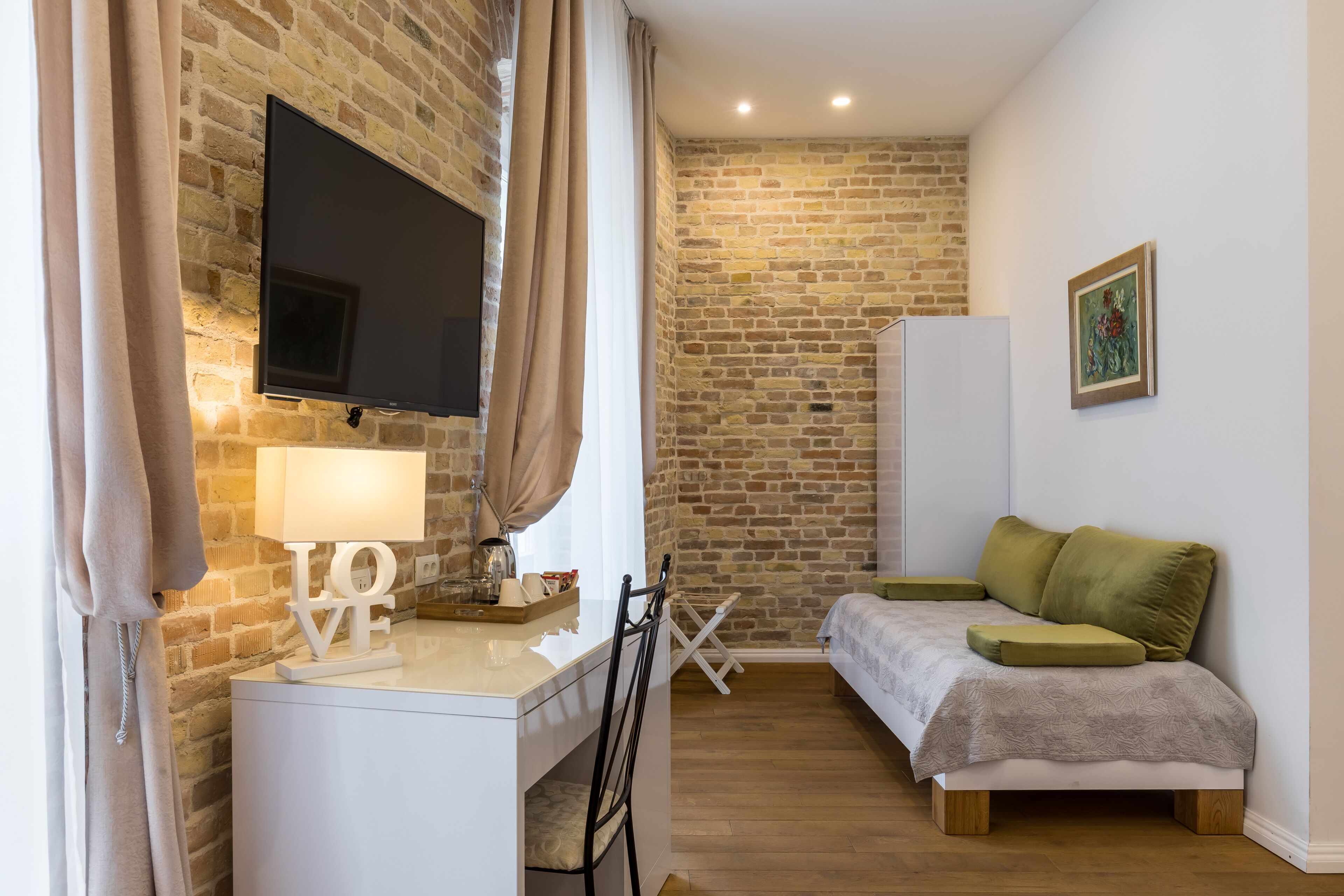 A cozy room with a brick wall, a flat-screen TV, a desk with a lamp, a bed with green pillows, and a small table with a tray.