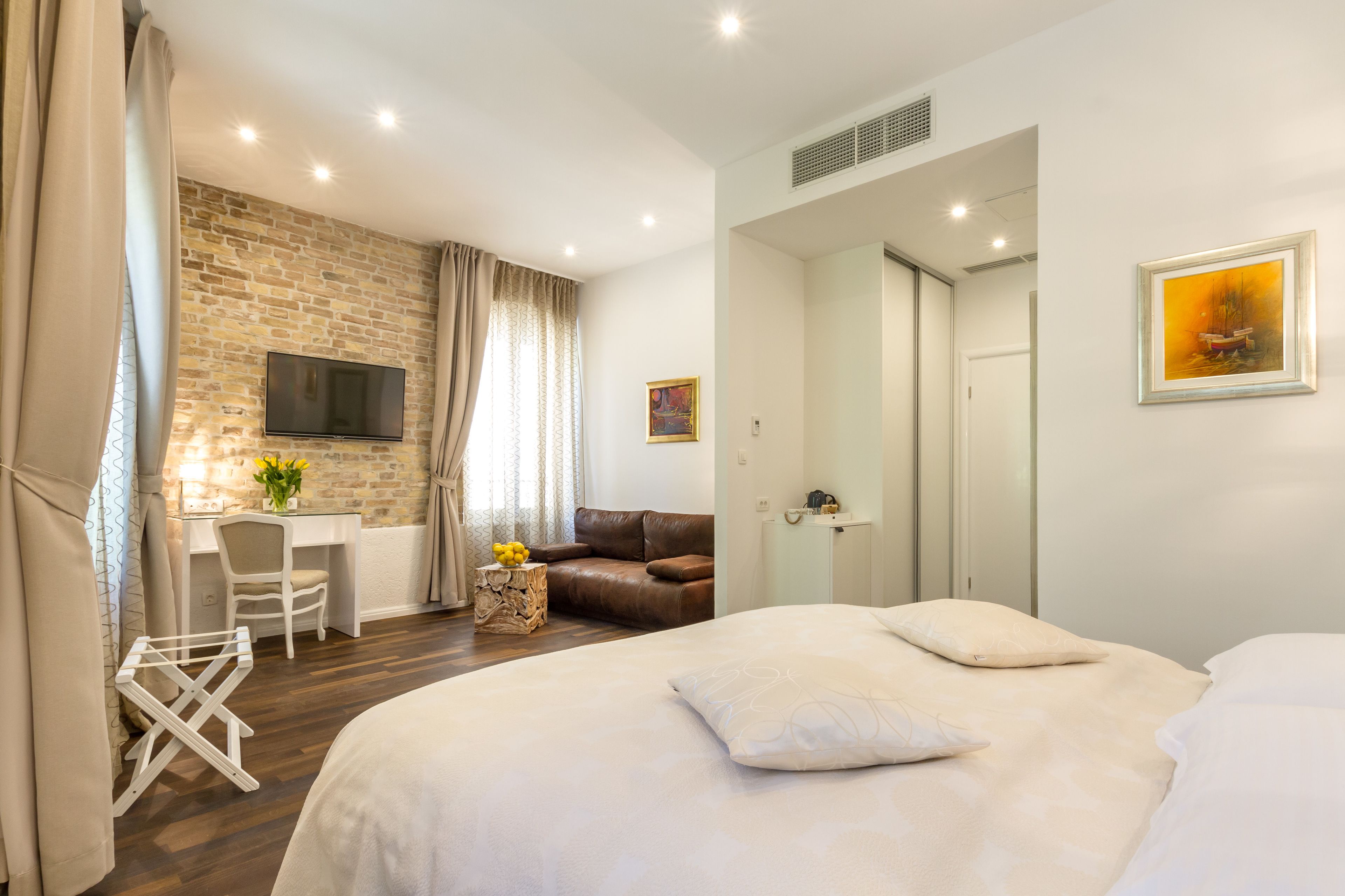 A modern hotel room with a brick wall, a flat-screen TV, a desk, a brown leather sofa, a bed with white bedding, and a painting on the wall.