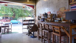 A cafe with a brick wall, wooden chairs, a counter with a computer, and a view of the street outside. - Ok Chic Phuket Hostel (Phuket)